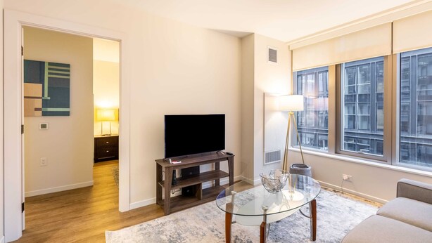 A living room with a flat-screen TV on a wooden stand, a glass coffee table, a sofa, and a large window with a city view. - Modern Apt with Gym Near Restaurants (Boston, MA)