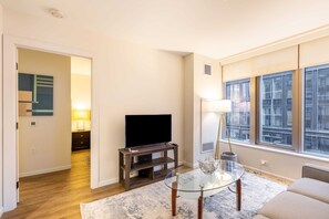 A living room with a flat-screen TV on a wooden stand, a glass coffee table, a sofa, and a large window with a city view. - Modern Apt with Gym Near Restaurants (Boston, MA)