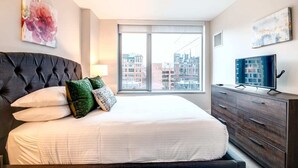 A modern bedroom with a large bed, a dresser, a television, and a city view through the window.