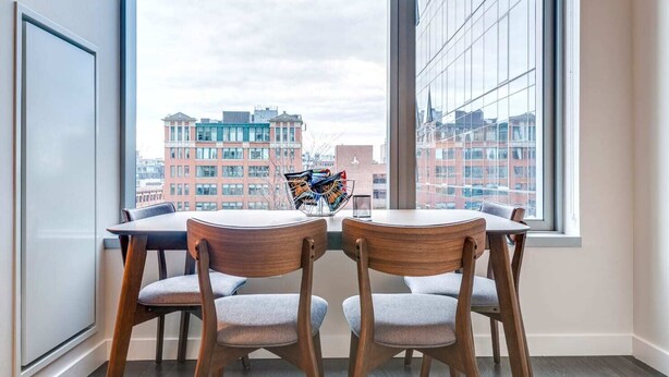 A dining area with two wooden chairs and a table, a window with a view of buildings, and a magazine rack with magazines.
