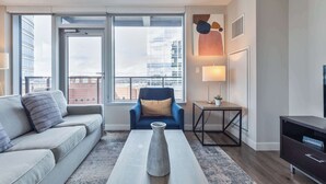 A modern living room with a sofa, a coffee table, a chair, a TV stand, and a large window with a city view.