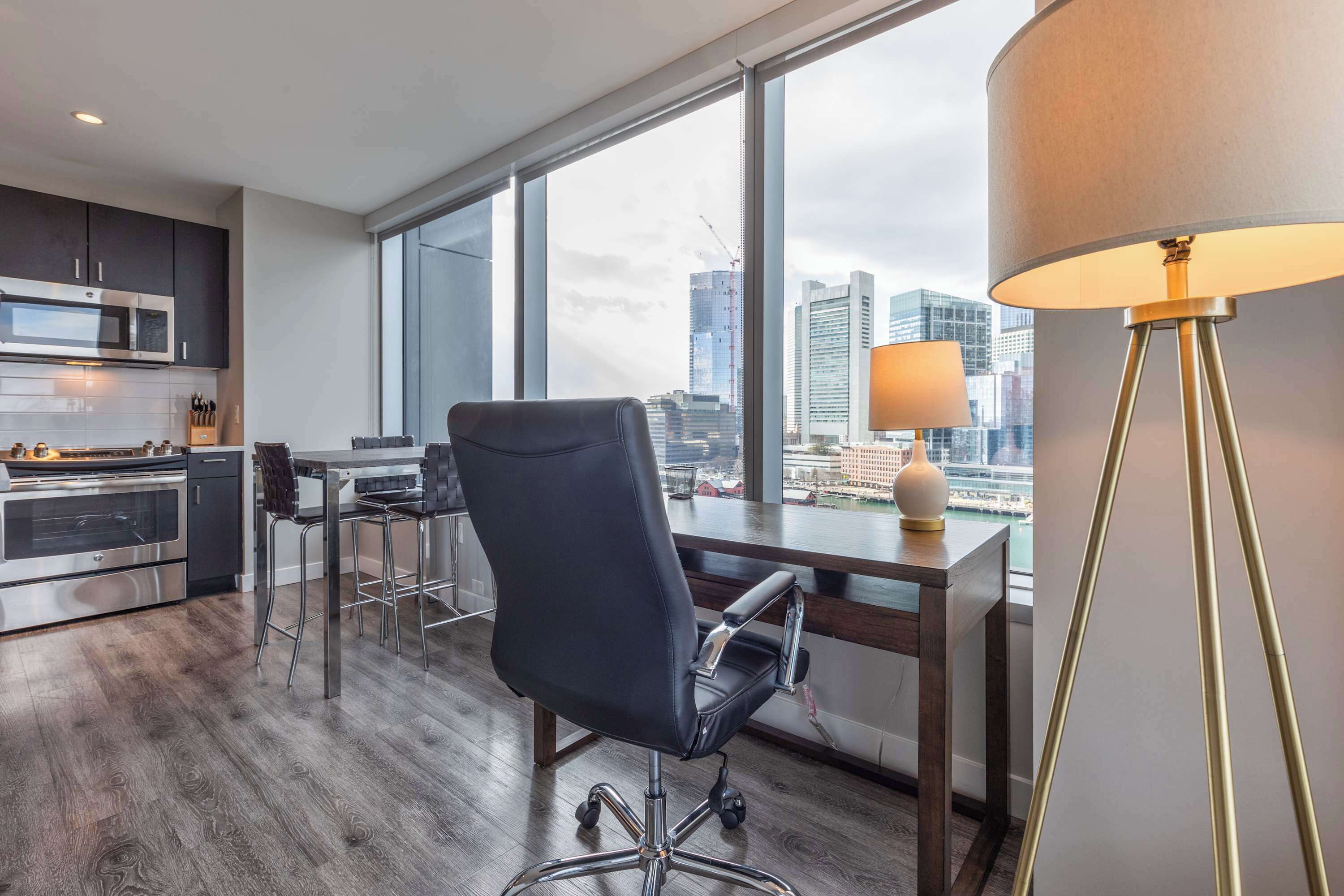 A modern office space with a large window, a desk, a chair, a lamp, and a kitchenette.