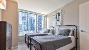 A bedroom with two beds, a dresser, a window with a city view, and a painting on the wall.