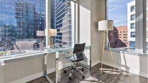 A modern office space with a glass desk, a black office chair, a floor lamp, and large windows offering a city view.
