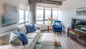 Ein modernes Wohnzimmer mit einer Couch, einem Hocker und einem blauen Sessel. Ein Fernseher steht auf einem Holzkonsole und eine Lampe auf einem kleinen Tisch.