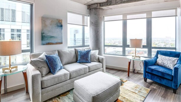 Ein modernes Wohnzimmer mit grauem Sofa, blauem Sessel und Blick auf die Stadt durch große Fenster.