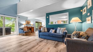 A spacious living room with a blue sofa, a wooden coffee table, and a large mirror on the wall. There is a sliding glass door leading to a balcony with a hot tub.