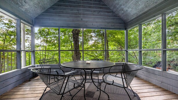 A covered outdoor seating area with a round table and chairs, large windows, and a view of trees.