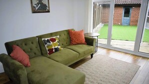 Un canapé vert agrémenté d’un coussin à motifs et d’un coussin rouge, une petite table ronde et, à travers une porte vitrée, une vue sur une pelouse verdoyante.