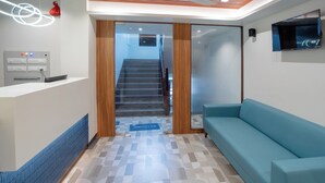 A reception area with a reception desk, a blue sofa, a patterned floor, and a staircase leading to an upper floor. - Hotel Anugraha NX (Palghar)
