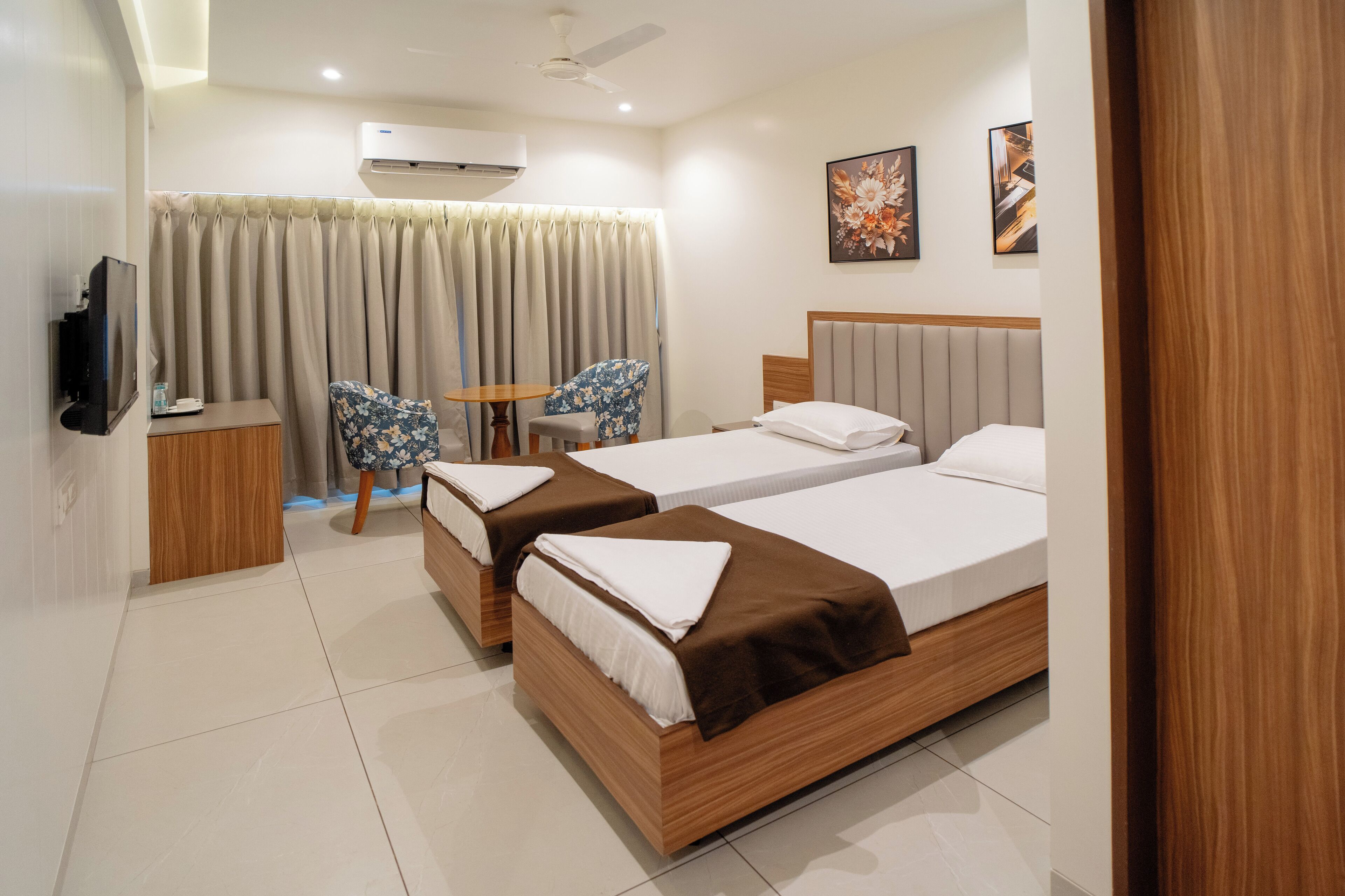 A hotel room with two beds, a television, a small table, a chair, and a window with curtains.