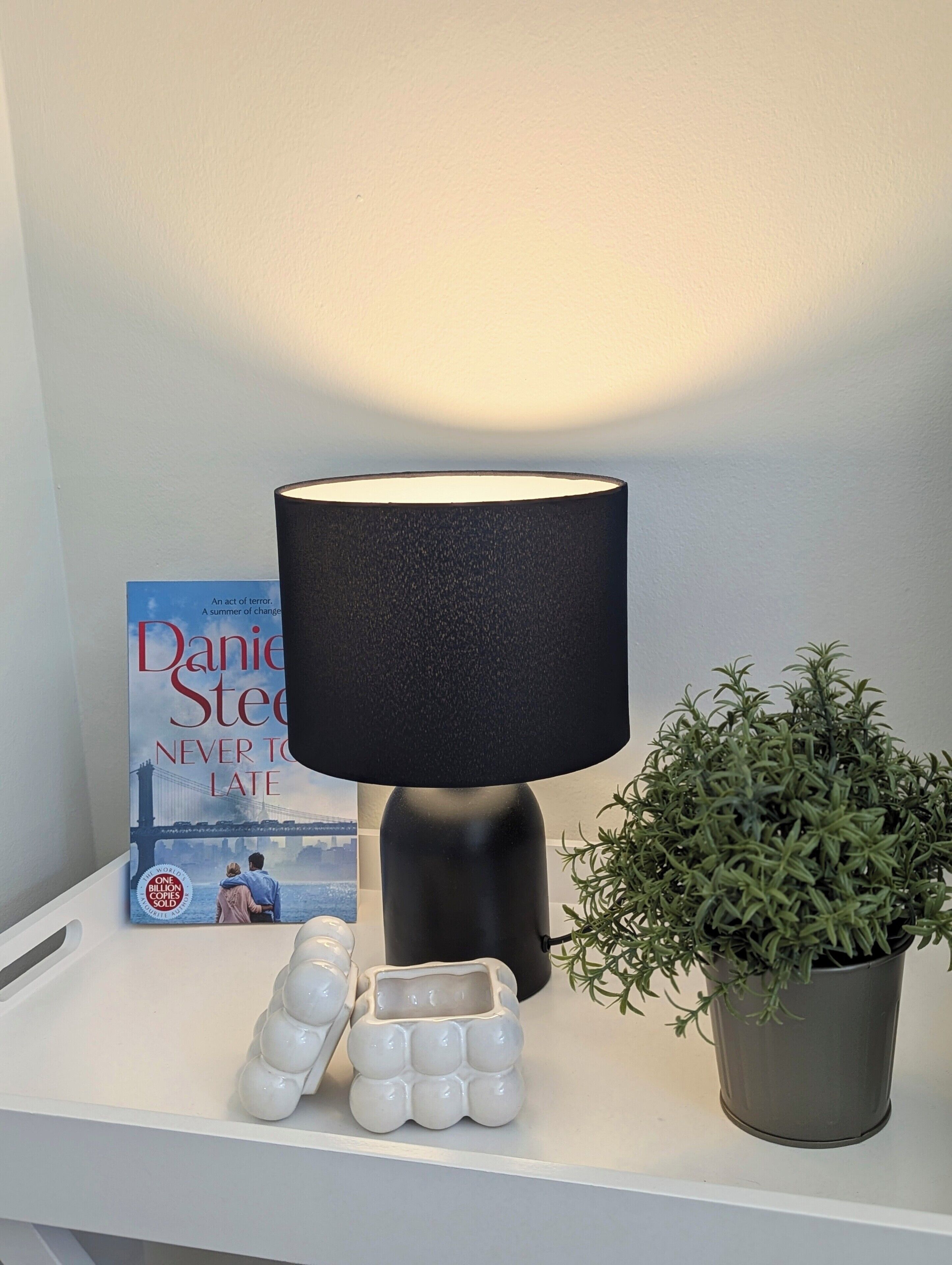 A bedside table with a black lamp, a book titled 'Never to Late', a small potted plant, and decorative eggs.