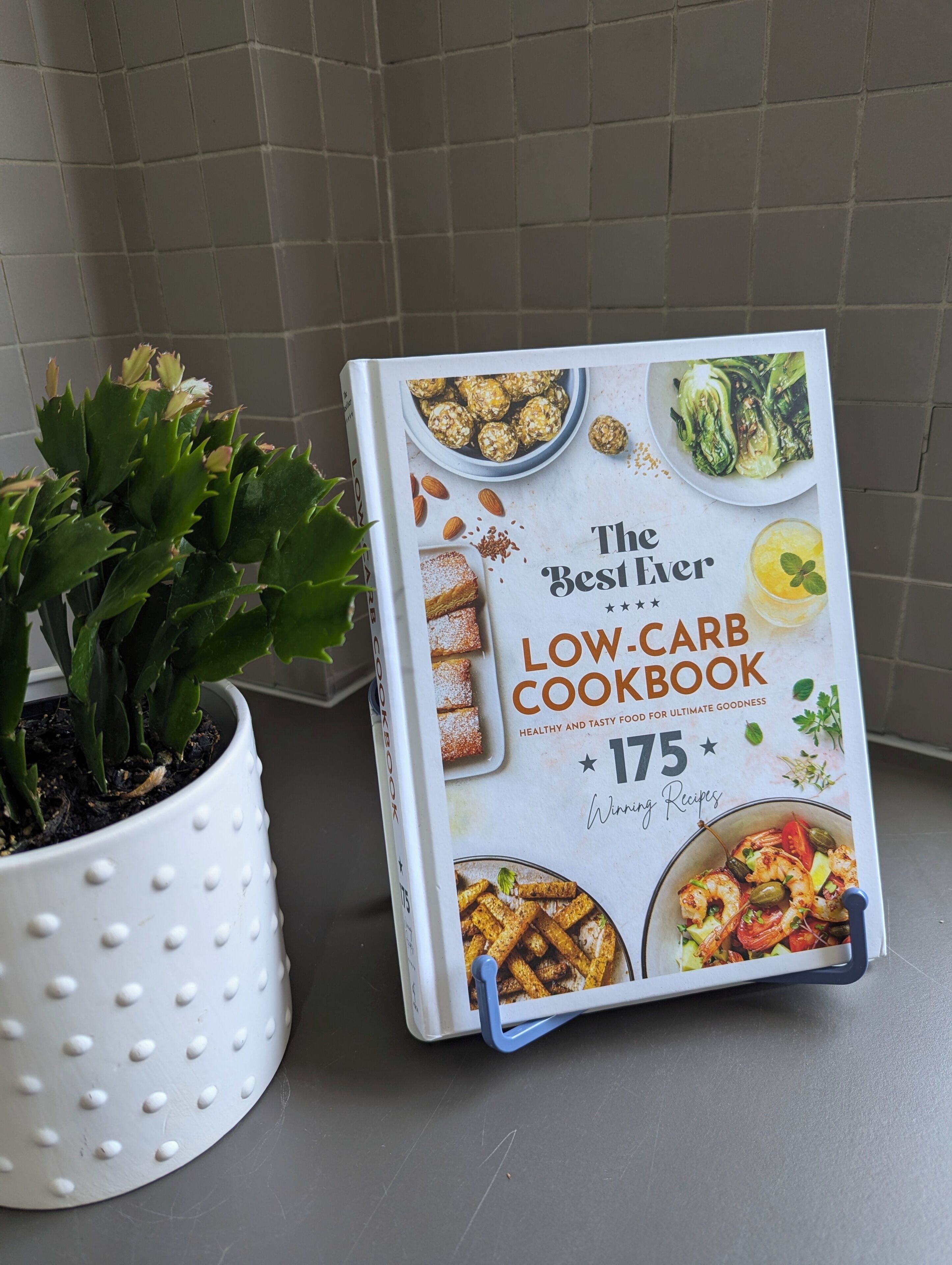 Un libro di cucina intitolato "The Best Ever Low-Carb Cookbook" con 175 ricette vincenti, posizionato accanto a una pianta in vaso su un piano di lavoro in cucina.
