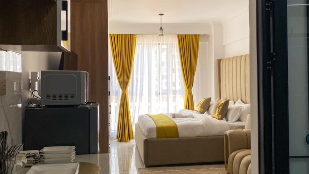 A modern hotel room with a bed, a desk with a computer, and a kitchen area. - Smart Homes - The Luxe Retreat (Nairobi)