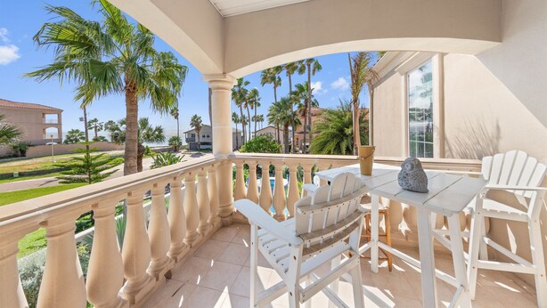 A balcony with white chairs and a table, overlooking a pool and palm trees.