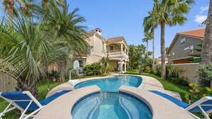 A swimming pool with lounge chairs, surrounded by palm trees and a large house with a balcony.