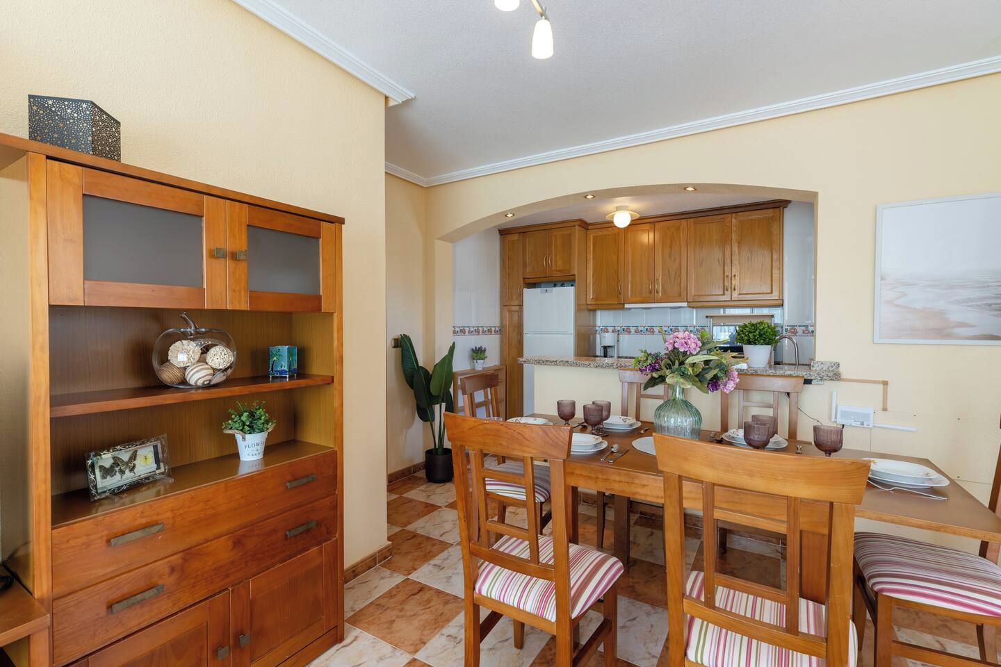 A dining area with wooden furniture, a kitchen in the background, and a checkered floor.