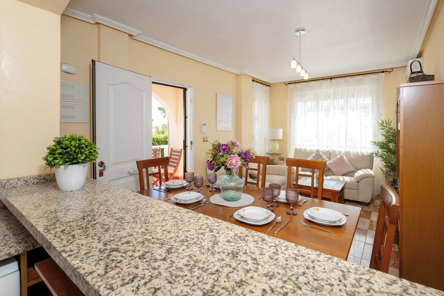 Un comedor con encimera de granito, sillas de madera, una mesa dispuesta para cuatro personas y un jarrón con flores.