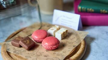 A tray with macarons and chocolates on a wooden board, a coffee cup, and books in the background.