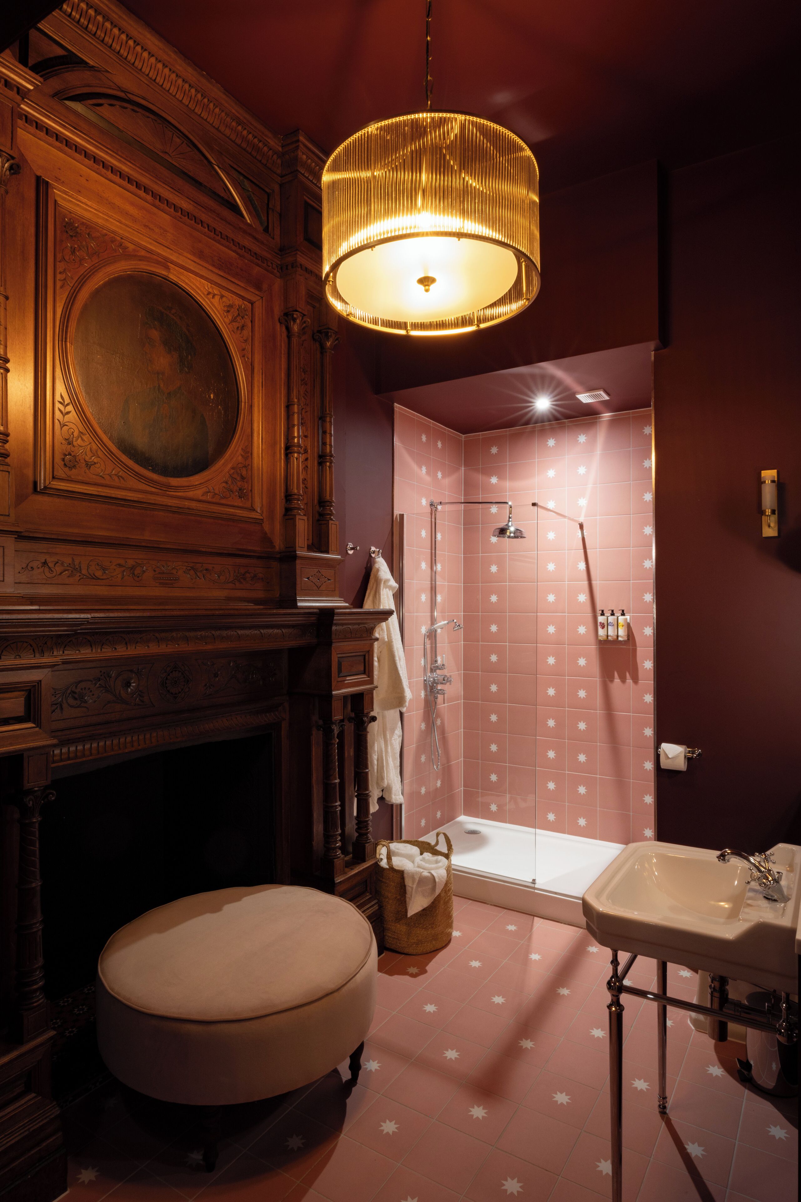 A bathroom with a wooden vanity, a round stool, a shower with a glass door, and a hanging light fixture.