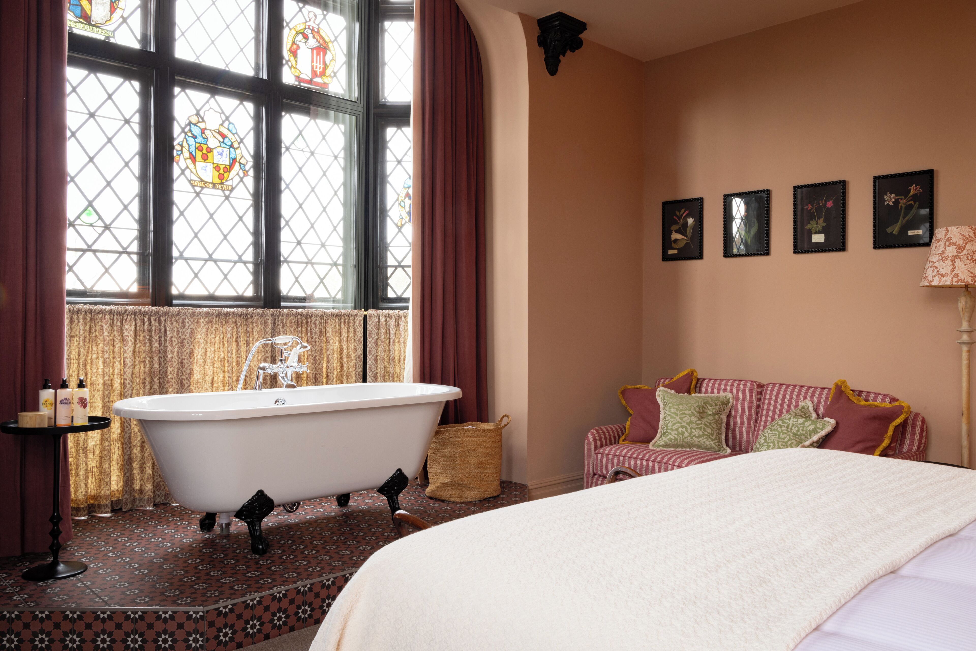 Una habitación con una bañera tipo pata de león, una cama, una mesita con artículos de aseo y una ventana con vidrieras.