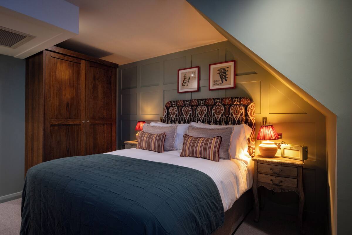 A bedroom with a large bed, a wooden wardrobe, a nightstand with a lamp, and two framed pictures on the wall.
