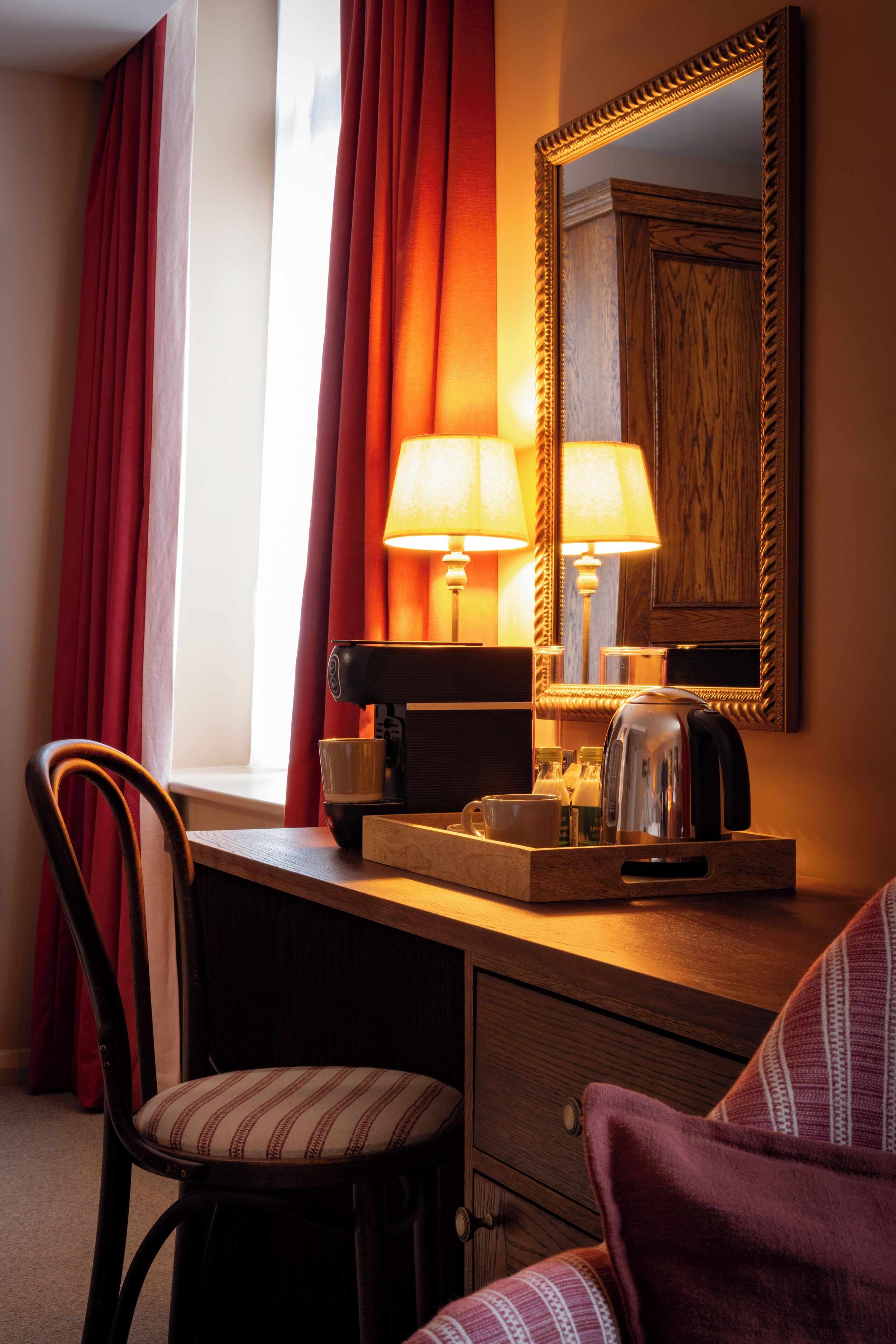 A hotel room with a wooden desk, a chair, a lamp, a kettle, and a mirror.