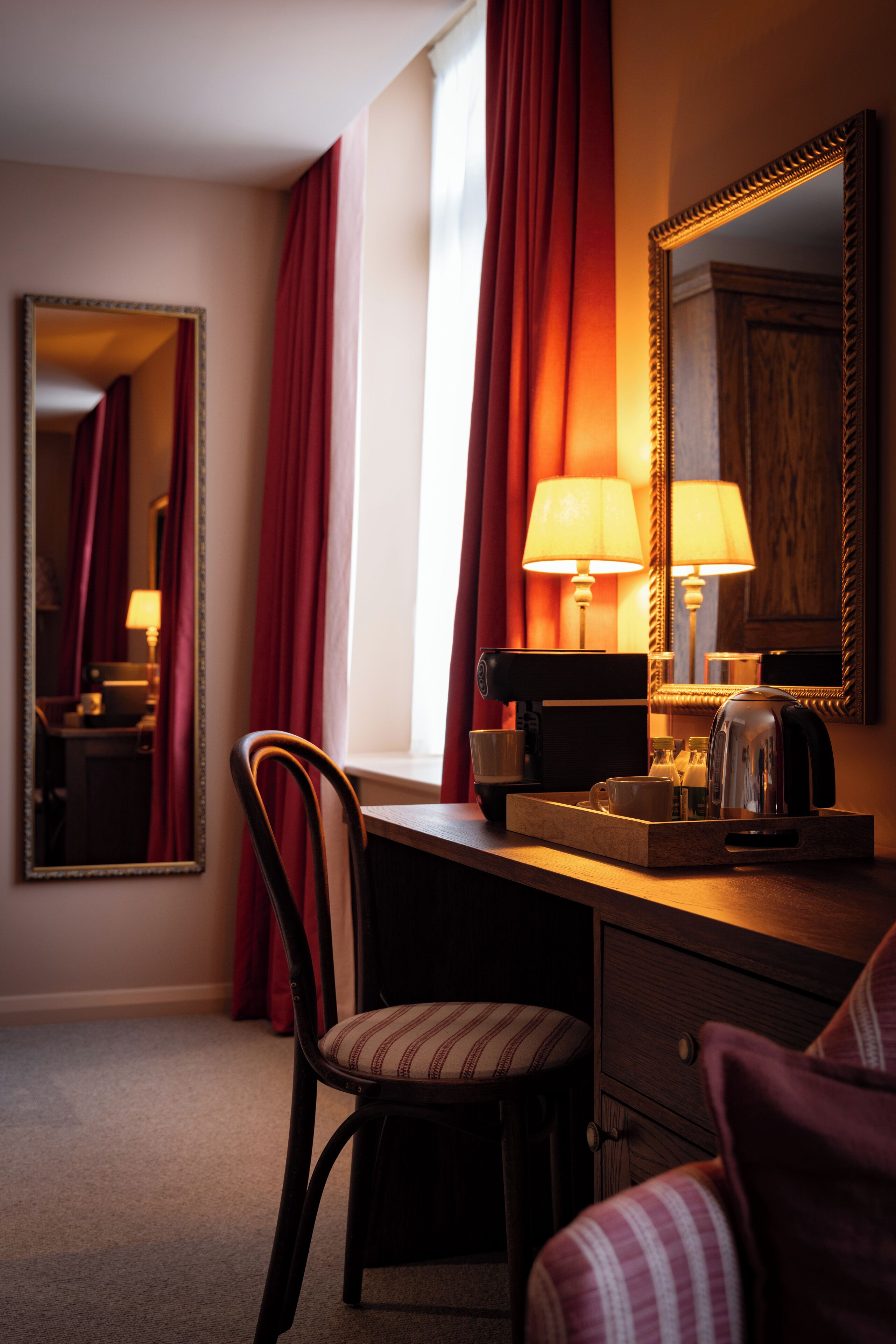 A hotel room with a wooden desk, a chair, a mirror, and a window with red curtains.