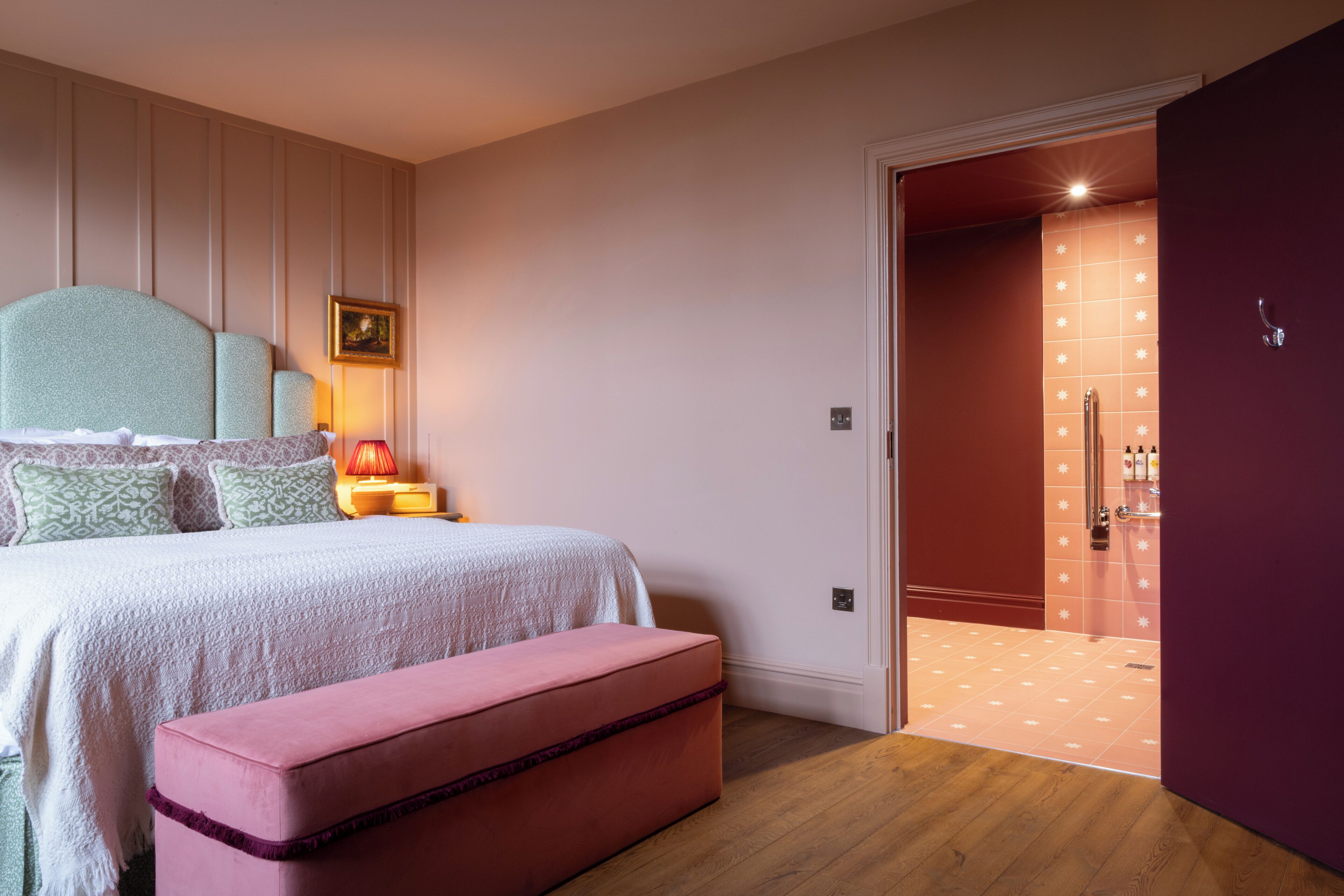 Ein Schlafzimmer mit einem Bett, einem Nachttisch, einer Nachttischlampe und einer pinkfarbenen Bank.