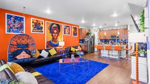 A modern living room with a blue rug, orange walls, and a kitchen area with wooden cabinets and a bar. - 420Friendly Game Arcade DancePole Airport DIA (Denver)