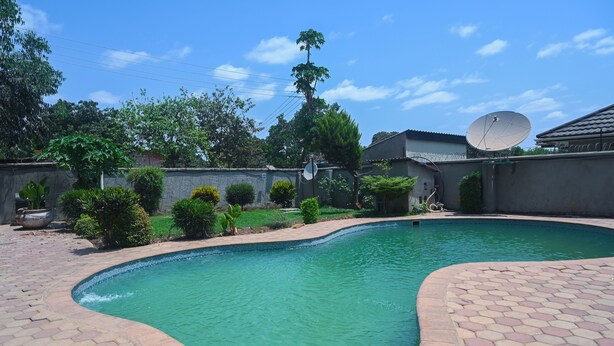 Uma piscina com borda de azulejos hexagonais, rodeada por um jardim com diversas plantas e uma antena parabólica.
