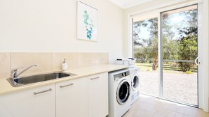 A modern laundry room with a washing machine, a sink, and a large window offering a view of a garden. - Our Multi-Award Winning Hawkesbury Holiday Home (East Kurrajong)