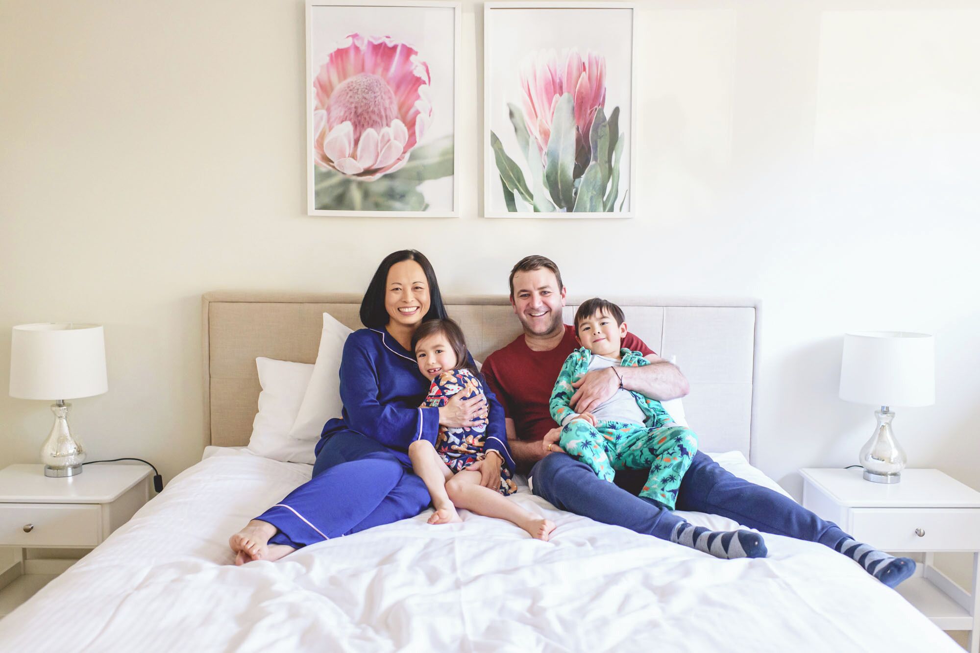 Une famille de quatre personnes est allongée sur un lit recouvert de draps blancs. Deux lampes sont fixées au mur et deux œuvres d’art florales encadrées décorent la pièce.