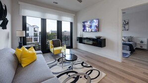 A modern living room with a sofa, a small table, a TV, and a balcony with a view of buildings. - West End Getaway for 5 w/Pool! (Nashville)