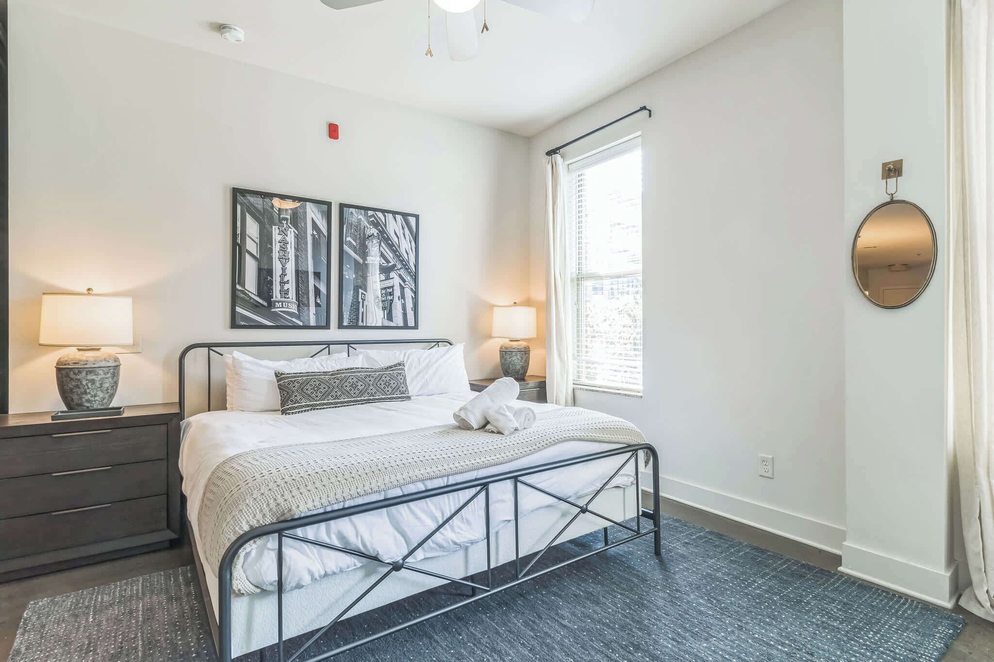 A neatly arranged bedroom with a bed, two bedside lamps, a wall-mounted mirror, and two framed pictures.