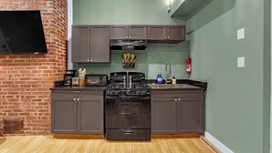A kitchen with a brick wall, a flat-screen TV, dark cabinets, a stove, and a microwave. - Lovely Little Loft-Style 2-Bedroom in Tulsa (Tulsa)