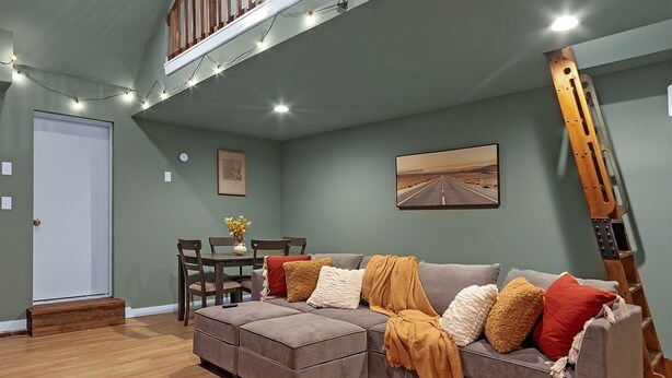 A spacious living area with a grey sectional sofa, wooden flooring, a dining table with chairs, a staircase with wooden railings, and string lights. - Lovely Little Loft-Style 2-Bedroom in Tulsa (Tulsa)