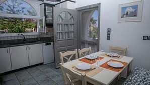 A dining area with a table set for four, a kitchen with white cabinets, and a view of greenery outside. - Villa Anna with Panoramic view (Santorini)