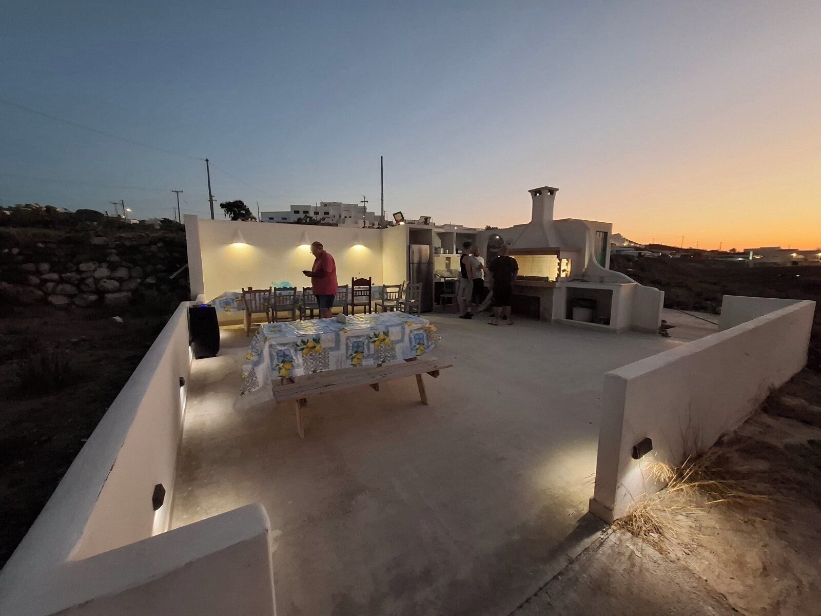 Un espace extérieur aménagé pour prendre les repas, avec une table dressée pour deux personnes, une personne debout près de la table et un bâtiment avec une cheminée en arrière-plan.