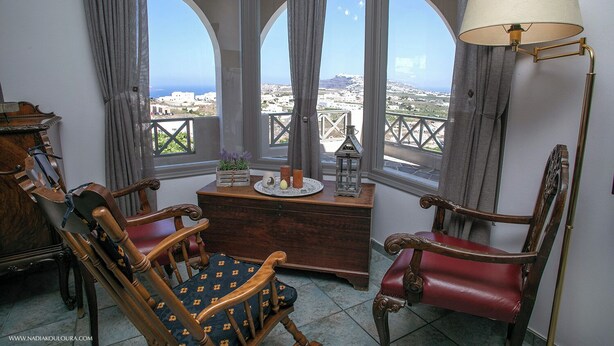 A room with a view of a coastal city, featuring two wooden chairs, a small wooden table with decorative items, and a floor lamp. - Villa Anna with Panoramic view (Santorini)