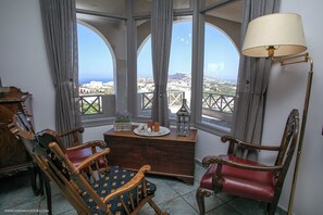 A room with a view of a coastal city, featuring two wooden chairs, a small wooden table with decorative items, and a floor lamp. - Villa Anna with Panoramic view (Santorini)