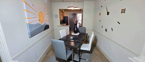 A dining room with a table, chairs, a clock, and a sun painting on the wall.