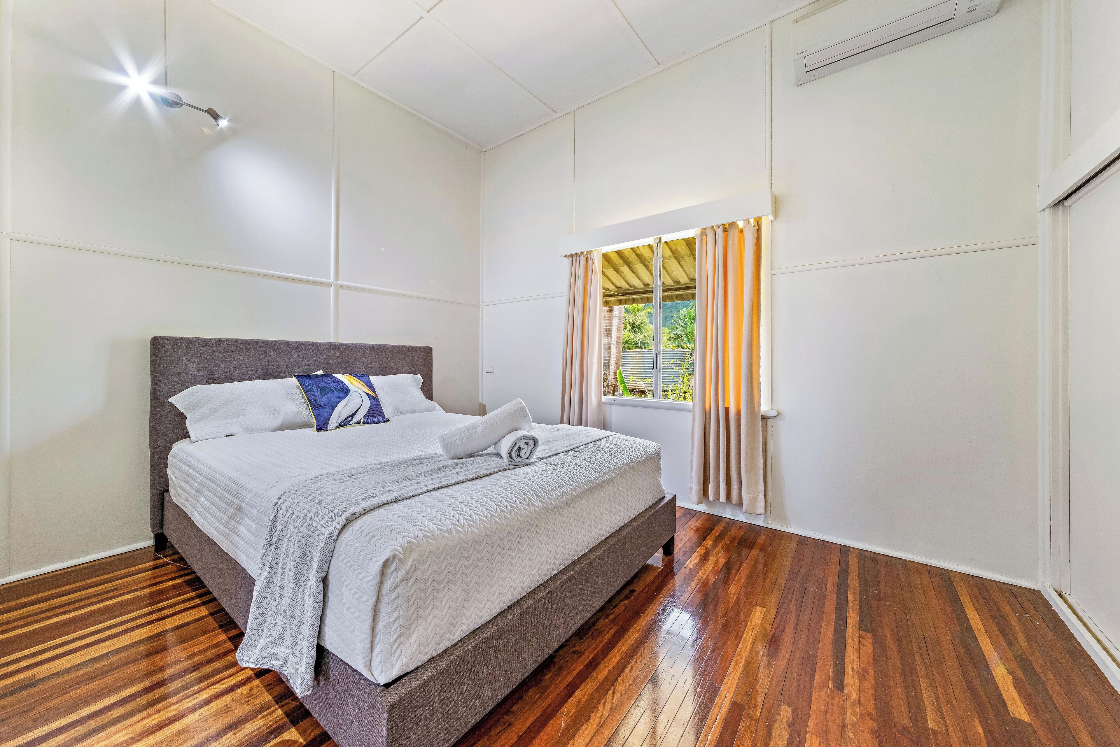 Ein Schlafzimmer mit einem großen Bett, Holzböden, einer Deckenleuchte und einem Fenster mit Vorhängen.