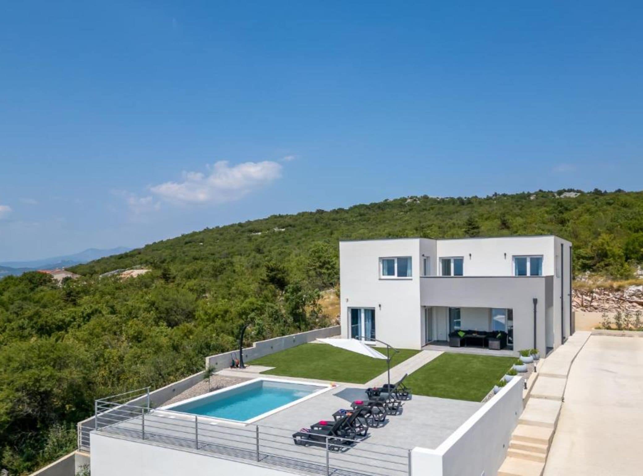 A modern two-story house with a swimming pool, a patio with lounge chairs, and a view of a green hillside.