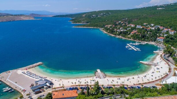 Vue aérienne d’une ville côtière avec une plage, une mer d’un bleu limpide et des collines environnantes.