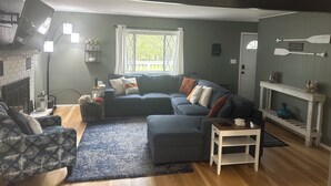 Een gezellige woonkamer met een donkerblauwe bank, een fauteuil met een patroon en een witte bijzettafel. Een open haard met bovenblad en wandlampen is zichtbaar.