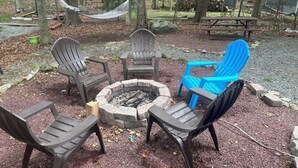 Een terras in het groen met Adirondack stoelen, een vuurplaats en een hangmat.
