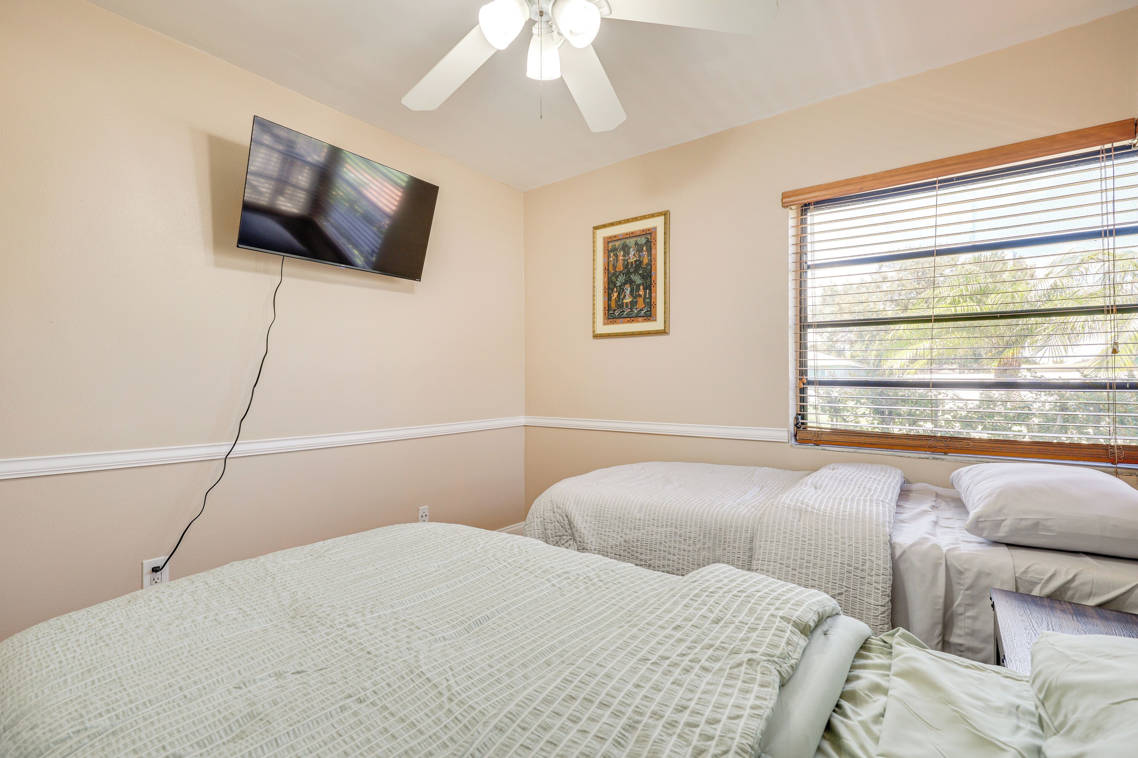 A bedroom with two beds, a ceiling fan, a TV mounted on the wall, a window with blinds, and a framed picture.