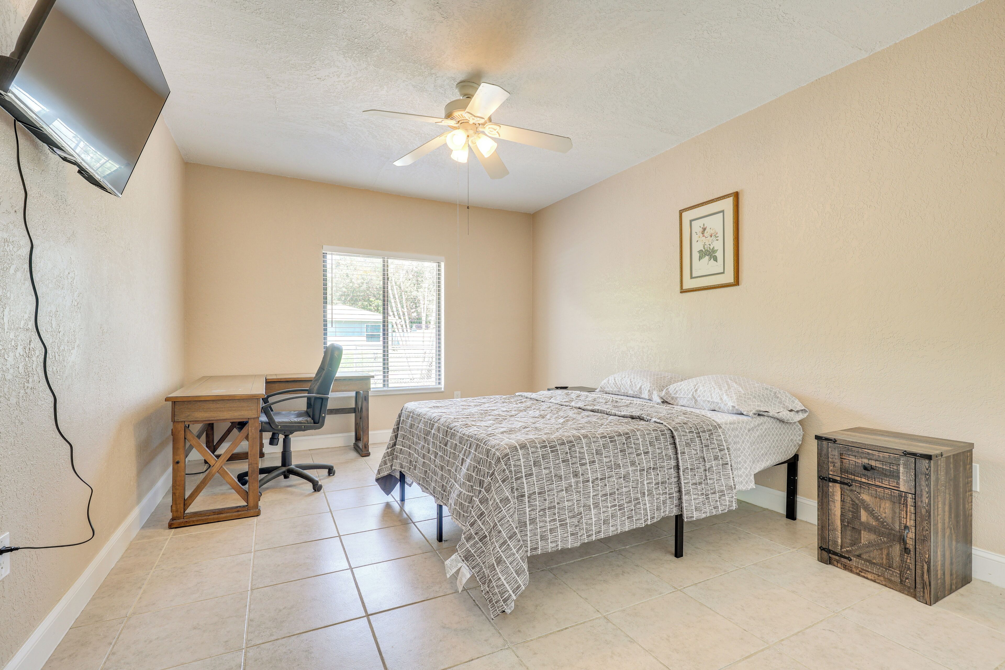 A bedroom with a bed, a desk, a chair, a ceiling fan, a TV mounted on the wall, and a window.