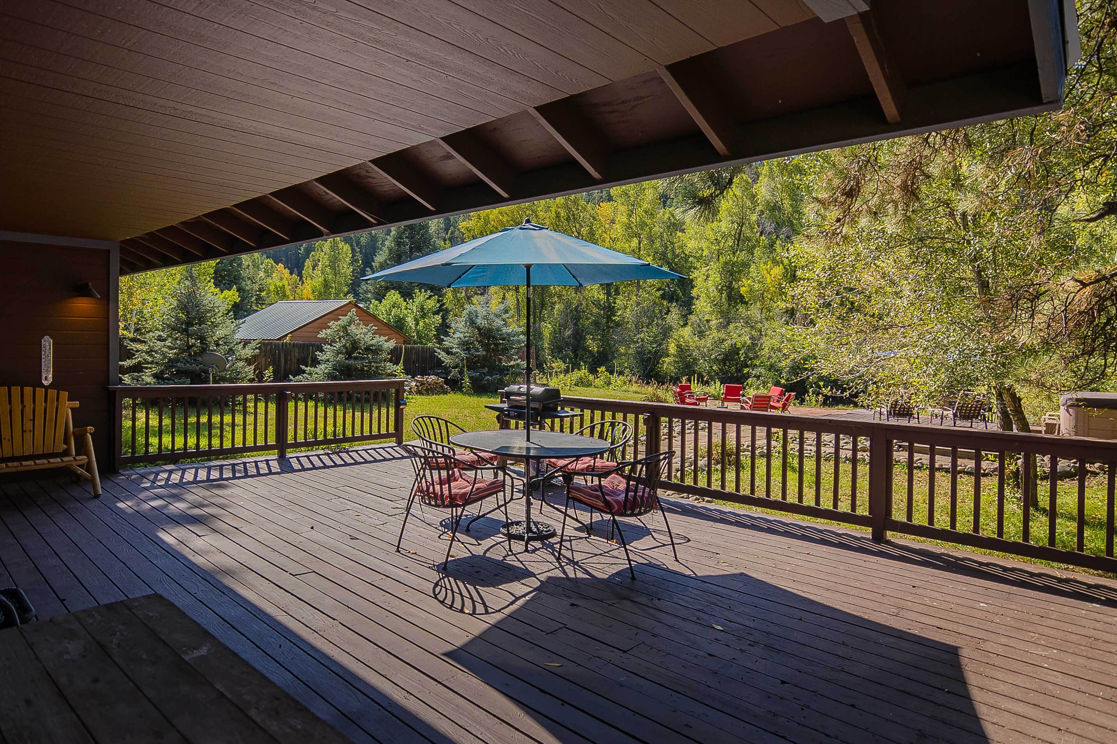 A wooden deck with a table and chairs, a blue umbrella, and a view of a forested area.